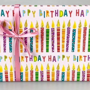 Puede incluir: Una caja de regalo blanca con una cinta rosa. La caja est&aacute; envuelta en papel con un patr&oacute;n de velas de colores y las palabras "Feliz cumplea&ntilde;os".
