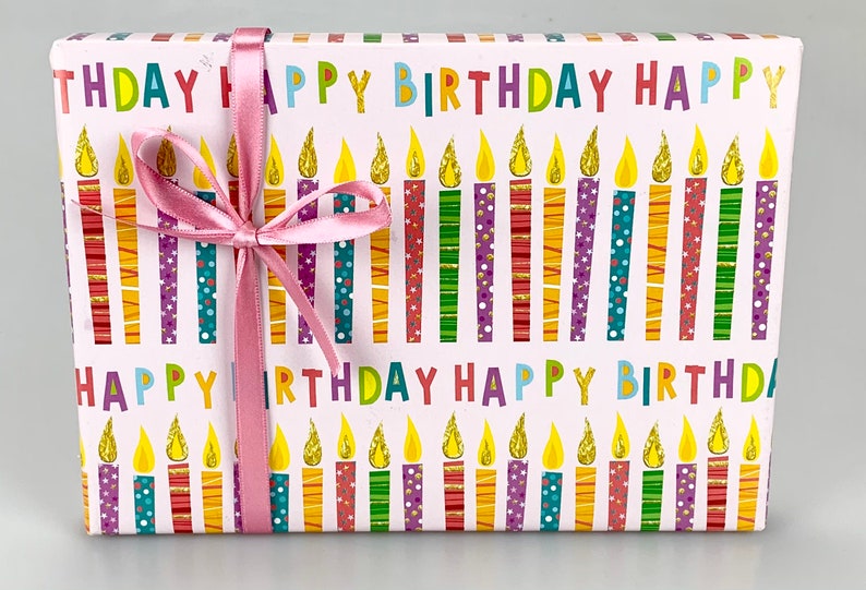 Puede incluir: Una caja de regalo blanca con una cinta rosa. La caja est&aacute; envuelta en papel con un patr&oacute;n de velas de colores y las palabras "Feliz cumplea&ntilde;os".