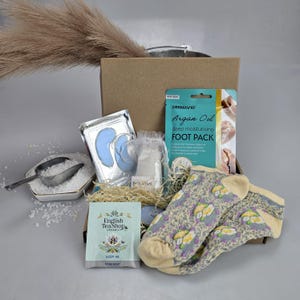 Può includere: Una scatola regalo piena di articoli per la cura di sé, tra cui un paio di calzini, una maschera per i piedi, maschere per gli occhi, sali da bagno, una bustina di tè e una candela. La scatola è piena di carta triturata e ha un esterno in cartone marrone.
