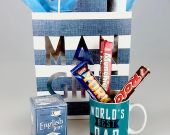 May include: A gift bag with blue and white stripes and the word "MA" cut out. Inside are candy bars, a mug that says "World's Best Dad", and a box of English tea. Blue tissue paper is visible.