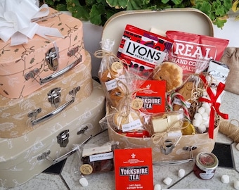 May include: A gift hamper set featuring three stacked cardboard suitcases in light pink and beige with white botanical prints, topped with a white bow. The open suitcase contains assorted food items including tea bags, cookies, and a Merry Christmas tag.