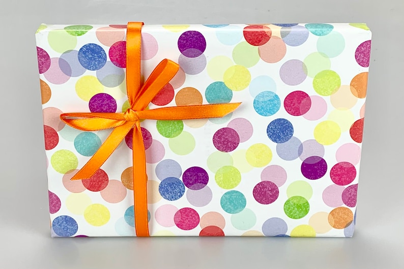 Puede incluir: Una caja de regalo blanca con un patr&oacute;n de lunares de colores y una cinta naranja atada en un lazo.