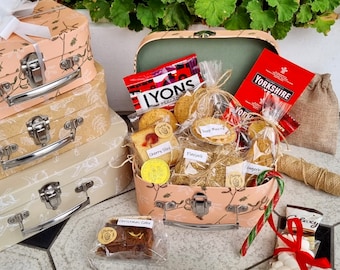 May include: A stack of decorative, peach-colored suitcases with silver hardware, topped with a white bow. The open suitcase reveals a selection of treats, including cookies and candies. A tag reads "merry Christmas."