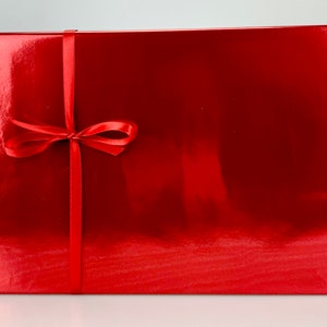 May include: A rectangular, glossy red gift box tied with a matching red satin ribbon and bow. The box has a reflective surface, suggesting a metallic or lacquered finish. The ribbon is centered vertically and tied in a neat bow.