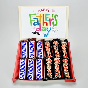 Könnte beinhalten: Eine beige Geschenkbox mit der Aufschrift "Happy Father's Day", einem Schnurrbart und einer Fliegenillustration. Die Box enthält Snickers- und Mars-Schokoriegel, angeordnet auf rotem Seidenpapier.
