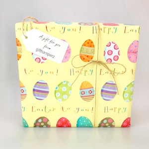 Puede incluir: Una caja de regalo amarilla con un lazo de cuerda marr&oacute;n. La caja est&aacute; envuelta en papel con un patr&oacute;n de huevos de Pascua coloridos y el texto "&iexcl;Feliz Pascua para ti!" Una etiqueta de regalo blanca est&aacute; unida a la caja con el texto "Un regalo para ti de gifthamperz".