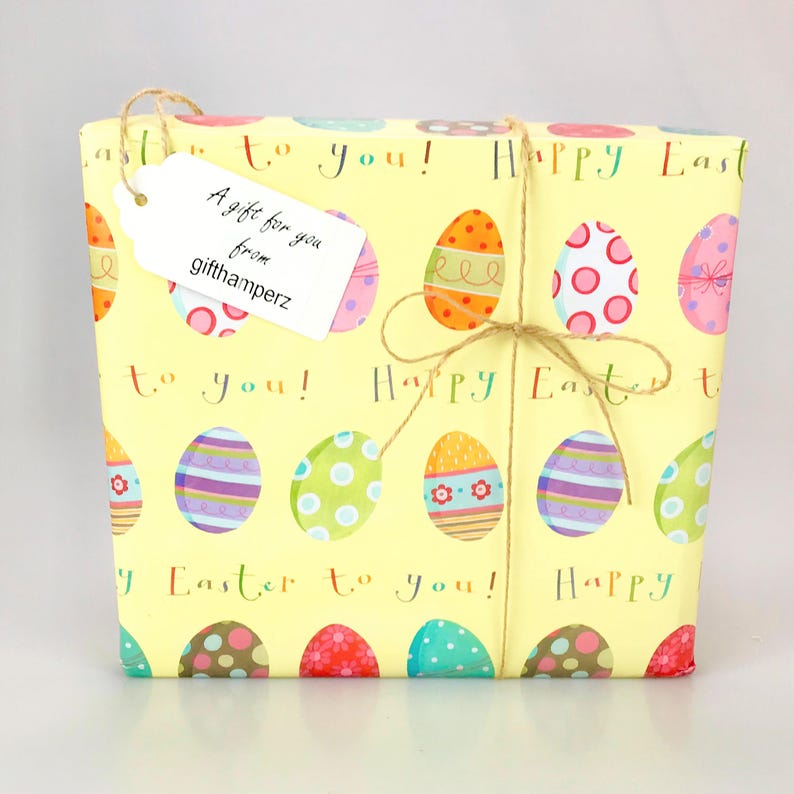 Puede incluir: Una caja de regalo amarilla con un lazo de cuerda marr&oacute;n. La caja est&aacute; envuelta en papel con un patr&oacute;n de huevos de Pascua coloridos y el texto "&iexcl;Feliz Pascua para ti!" Una etiqueta de regalo blanca est&aacute; unida a la caja con el texto "Un regalo para ti de gifthamperz".