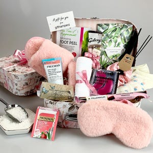 May include: A pink and white gift basket filled with self-care items, including a pair of fluffy pink slippers, a pink eye mask, a white towel, a pink satin scrunchie, a tea tree and peppermint foot peel, a Freeman anti-stress clay mask, a pack of under eye patches, a pack of tea, a small scoop of bath salts, and a box of dark chocolate tiramisu.