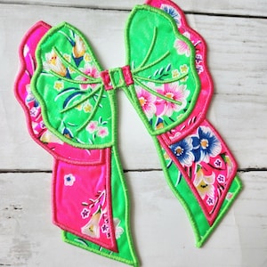 May include: A large, embroidered bow in vibrant colors. The bow features a bright green base with a pink outline and accents. The bow's center and tails showcase floral patterns in pink, blue, and yellow. The design is detailed with stitched lines.