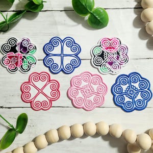May include: Six embroidered coasters with a clover design. The coasters are white with blue, pink, and red embroidery. One coaster has a floral design with pink, green, and blue embroidery.