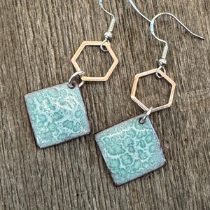 May include: A pair of earrings with a light blue square ceramic charm and a gold hexagon charm hanging from a silver hook.