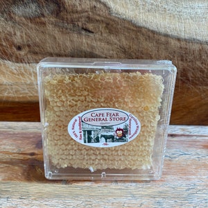 May include: A clear plastic container holds a honeycomb slab of raw and unfiltered honey. The label reads "Cape Fear General Store" and "Carolina Wildflower Honeycomb".