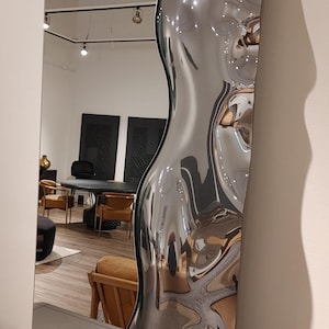 May include: A tall, rectangular floor mirror featuring a flat reflective surface on the left side and a three-dimensional, undulating, liquid-like metallic frame on the right side. The frame has a polished, chrome-like finish that reflects light.