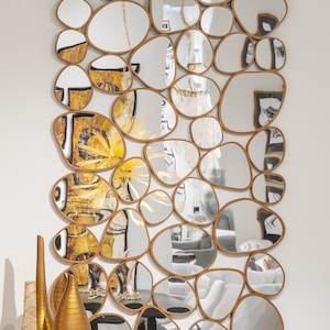 May include: A large, decorative wall mirror made of multiple, irregularly shaped, oval-shaped pieces of mirror glass. Each piece of mirror glass is framed in a thin, gold-coloured metal frame. The mirror is hanging on a white wall.