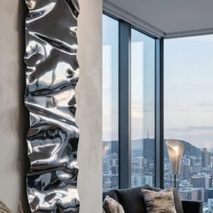 May include: A tall, vertical wall-mounted radiator with a highly reflective, polished chrome finish. The surface features a distinct, undulating, wave-like texture that creates distorted reflections of the surrounding room environment.
