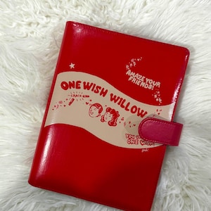 May include: A bright red notebook with a beige horizontal band across the center. The text reads ONE WISH WILLOW, AMAZE YOUR FRIENDS!, and YOU O ONE WISH!. It features illustrations of two faces, a wishbone, and stars. A red snap closure is on the right.