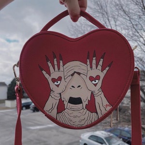 May include: Red heart-shaped purse with a hand-painted illustration of a creature with its eyes covered by hands with hearts on them. The creature has a dripping, melted appearance.
