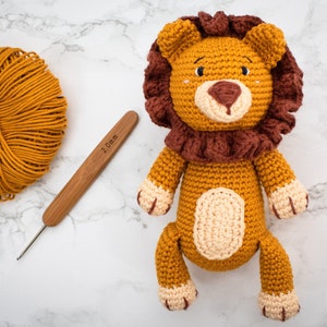 May include: A crocheted lion toy with a brown mane, yellow body, and cream-colored paws. The toy is sitting on a white marble surface with a ball of yellow yarn and a 2.0 mm crochet hook.