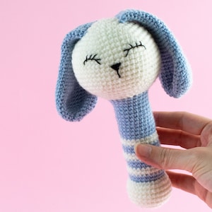 K&ouml;nnte beinhalten: Eine geh&auml;kelte Babyrassel mit einem blau-wei&szlig; gestreiften Griff und einem wei&szlig;en Hasenkopf mit blauen Ohren. Der Hase hat gestickte Augen und eine schwarze Nase.