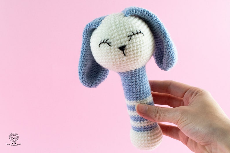 K&ouml;nnte beinhalten: Eine geh&auml;kelte Babyrassel mit einem blau-wei&szlig; gestreiften Griff und einem wei&szlig;en Hasenkopf mit blauen Ohren. Der Hase hat gestickte Augen und eine schwarze Nase.