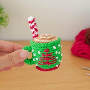 May include: A crocheted miniature mug of hot chocolate with a candy cane stirrer. The mug is green with white polka dots and a red Christmas tree design. The drink has a tan top and is held by a hand.