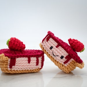 Puede incluir: Dos tartas de fresa de crochet con caras felices. Las tartas son rosas y rojas con una corteza marrón. Están sobre una superficie de mármol blanco.