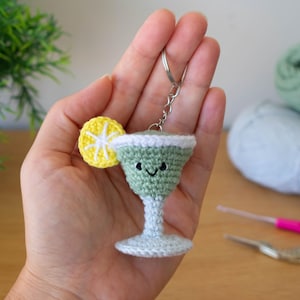 Puede incluir: Un llavero de ganchillo hecho a mano con forma de copa de cóctel con una rodaja de limón. La copa es de color verde claro con un borde blanco y una cara sonriente. La rodaja de limón es amarilla y blanca.