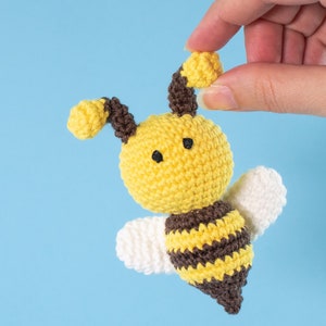 May include: A crocheted bee toy with yellow and brown stripes, white wings, and black eyes. The bee is held in a hand against a light blue background.