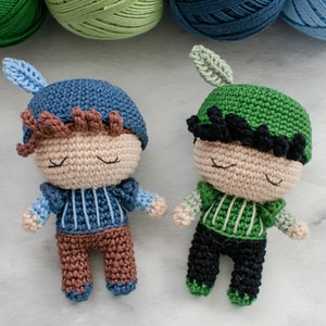May include: Two handmade crochet dolls, each with closed eyes, wearing hats and outfits. One doll has a blue hat and outfit, the other has a green hat and outfit. Yarn balls in shades of green and blue are in the background.