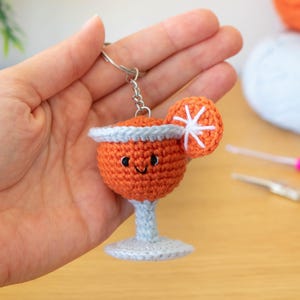 Puede incluir: Llavero de cóctel naranja tejido a ganchillo con una cara sonriente y una rodaja de cítricos blanca y naranja. La bebida tiene un borde y una base de color azul claro. El llavero se sostiene en una mano.