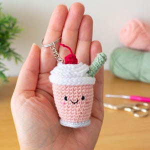 Puede incluir: Un llavero de ganchillo hecho a mano con forma de batido rosa con una cobertura de crema batida blanca, una cereza roja y una pajita verde. El llavero tiene una cara sonriente y está unido a un llavero plateado.
