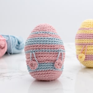 May include: Two crocheted Easter eggs, one pink and blue striped and one yellow and pink striped, with pink crocheted flowers on the bottom. Balls of yarn in pink, yellow, and blue are in the background.