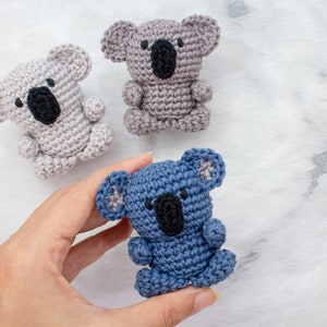 May include: Three crocheted koala bears, one grey, one light grey, and one blue. The blue koala is being held in a hand.