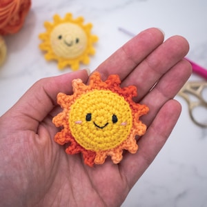 May include: A crocheted sun with a smiling face, orange and yellow yarn, and a black embroidered mouth. The sun is held in a person's hand.
