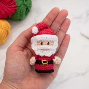 May include: A crocheted Santa Claus ornament in red and white with a white beard and black belt. The ornament is being held in a person's hand.