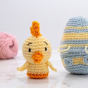 May include: A crocheted yellow chick with orange beak and crest, and a blue and yellow striped crocheted egg. The chick is standing on a white surface.