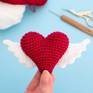 May include: A crocheted red heart with white wings. The heart is held in a hand against a light blue background.