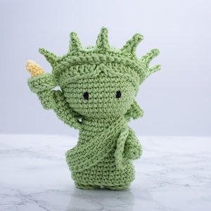May include: A green crocheted amigurumi doll of the Statue of Liberty holding a torch. The doll has black eyes and a yellow torch.