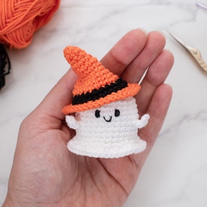 May include: A handmade crochet ghost wearing an orange and black witch hat. The ghost is white with a smiling face and small arms. The Halloween-themed decoration is held in a hand, with yarn and scissors in the background.
