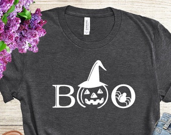 May include: Dark gray t-shirt with the word "BOO" in white, featuring a pumpkin with a witch hat and a spider in place of the "O". The shirt is paired with blue jeans and sandals, with a bouquet of purple flowers.