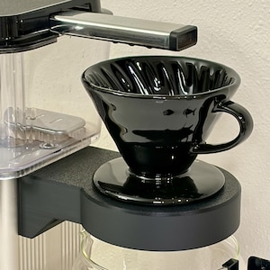 May include: A black ceramic pour-over coffee maker with a handle, sitting on a black base. The coffee maker is part of a larger silver and black coffee machine. The design features a ribbed interior and a glossy finish, perfect for brewing coffee.