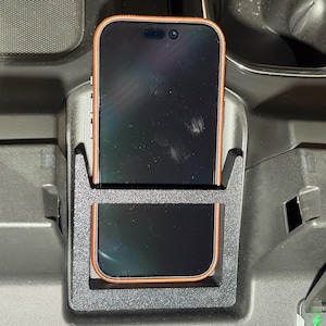 May include: A black phone holder with a phone inside. The phone has a black screen and a copper-colored frame. The phone holder is in a car's console.