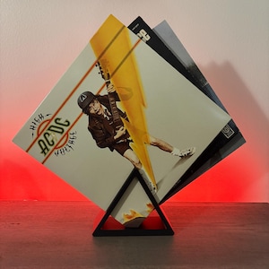 Pode incluir: Um suporte de exibição de discos de metal preto com três álbuns de vinil. A capa do álbum frontal apresenta a banda AC/DC com as palavras "High Voltage" e uma imagem de um músico tocando guitarra. O suporte está em uma superfície de madeira.