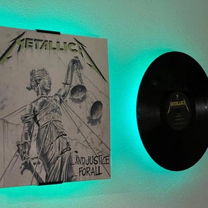 Peut inclure: Une pochette d'album encadrée de Metallica, "...And Justice for All", avec une illustration en niveaux de gris de Lady Justice. Un disque vinyle noir est monté à côté de la pochette. Le mur est éclairé par des lumières LED vertes.