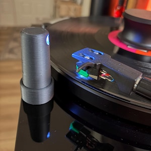 May include: A close-up of a black turntable with a vinyl record playing. The turntable features a black tonearm with a blue headshell and a stylus. A cylindrical, dark gray control knob is on the left side, with a blue light emanating from the top.