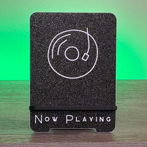 May include: Black phone stand with a textured surface and a white vinyl record graphic. The words "Now Playing" are printed in white at the bottom. A black rubber band holds the phone in place.