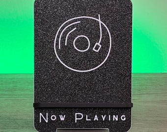 May include: Black phone stand with a textured surface and a white vinyl record graphic. The words "Now Playing" are printed in white at the bottom. A black rubber band holds the phone in place.