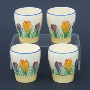 May include: Set of four cream-colored ceramic egg cups with a blue rim. Each cup features a hand-painted design of yellow, orange, and purple crocuses.