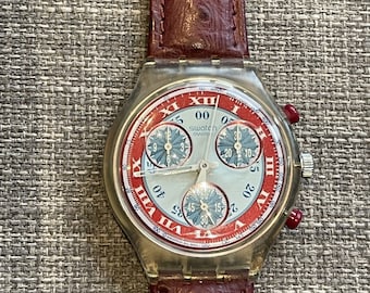 May include: A Swatch chronograph watch with a clear case, red and white dial, and a burgundy leather strap. The watch face features Roman numerals and three sub-dials. The strap is a textured, reddish-brown color.