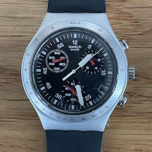 May include: A Swatch chronograph watch with a black dial, white hands, and a silver-colored case. The watch features a black rubber strap and multiple sub-dials. The watch face displays the text "Swatch SWISS".